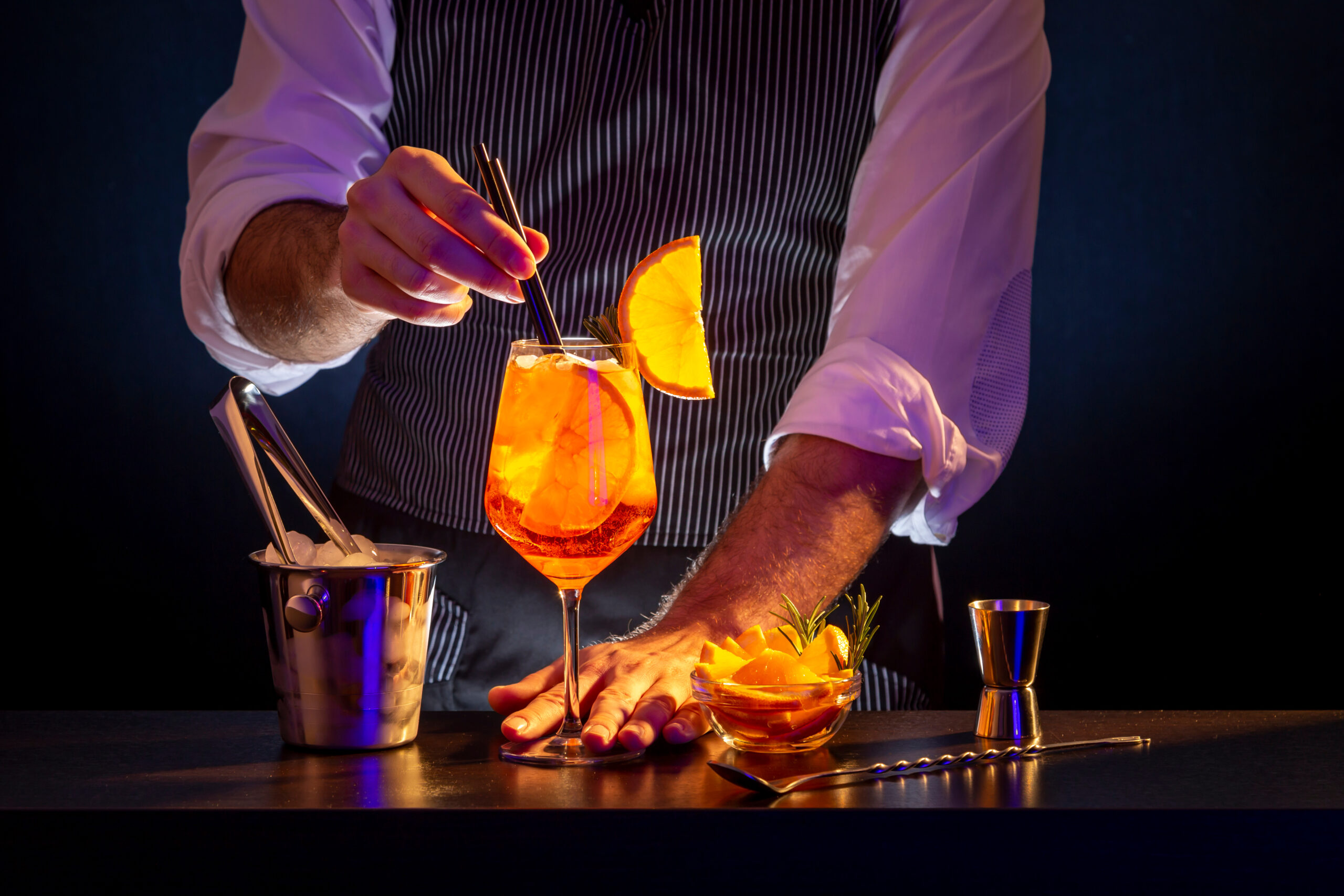 Bartender adding compostable drinking straws into Aperol spritz cocktail glass; barman serving orange liqueur cocktail in a wineglass on a bar counter.