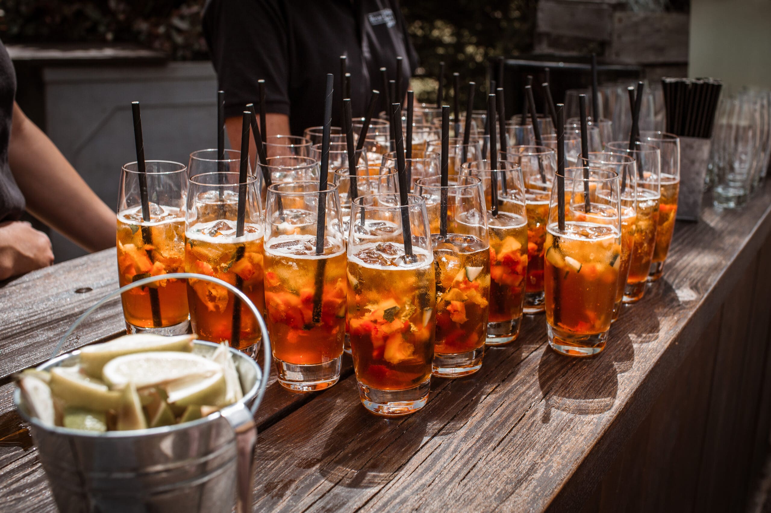 A row of beverages lined up on a rustic wooden bar. Each glass has a black, compostable, bioplastic straw in it. 